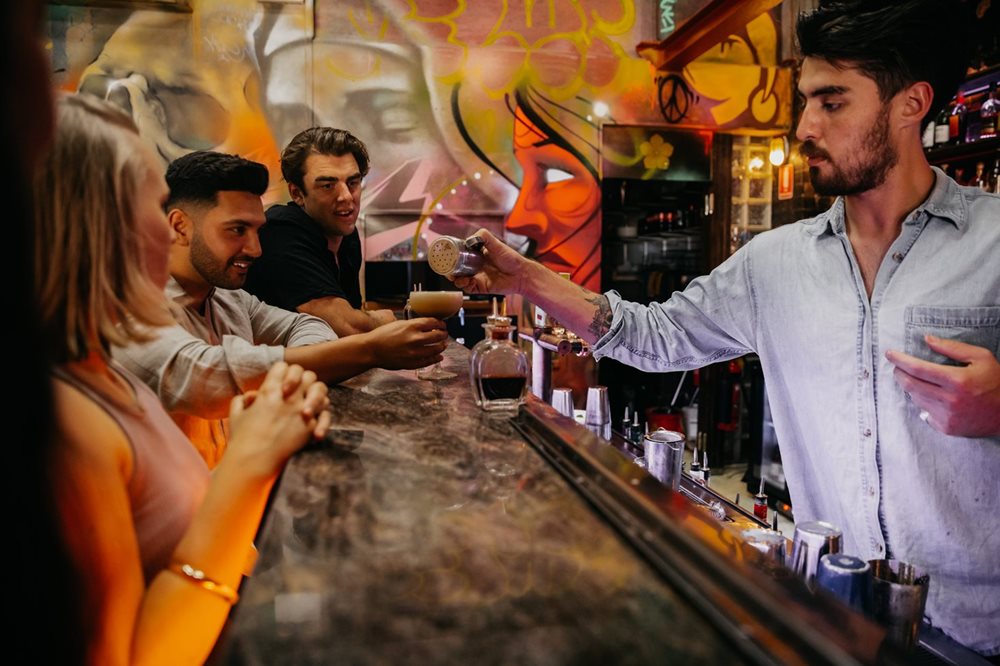 People sitting at a bar watching a mixologist serve cocktails