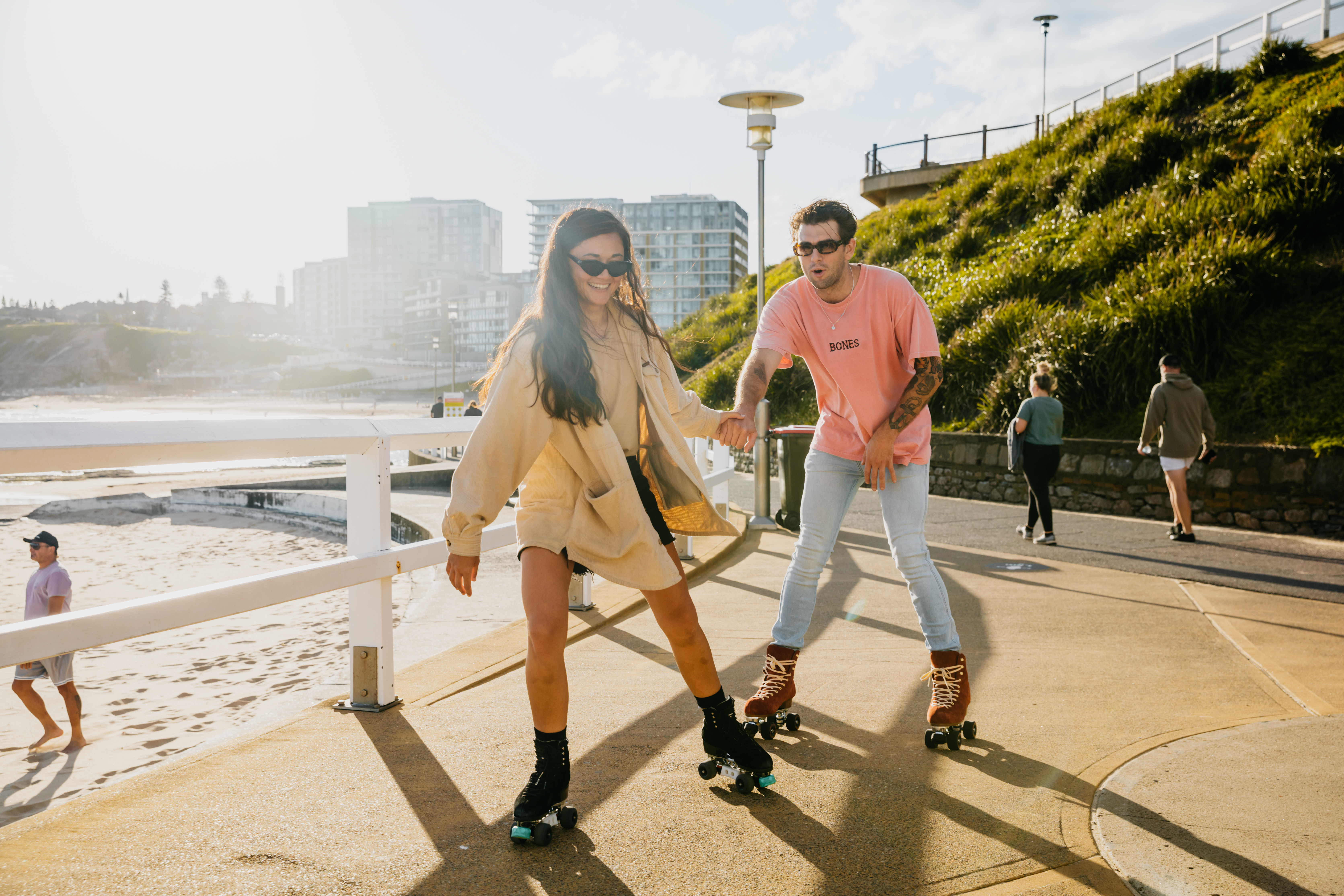 A couple roller skating on a path next to the beach with a city in the background